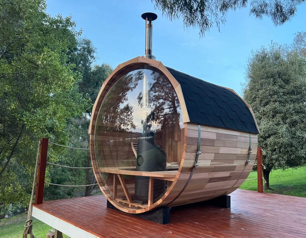 Cedar barrel sauna with panoramic glass end and wood heater flue on a deck in Cygnet, Tasmania