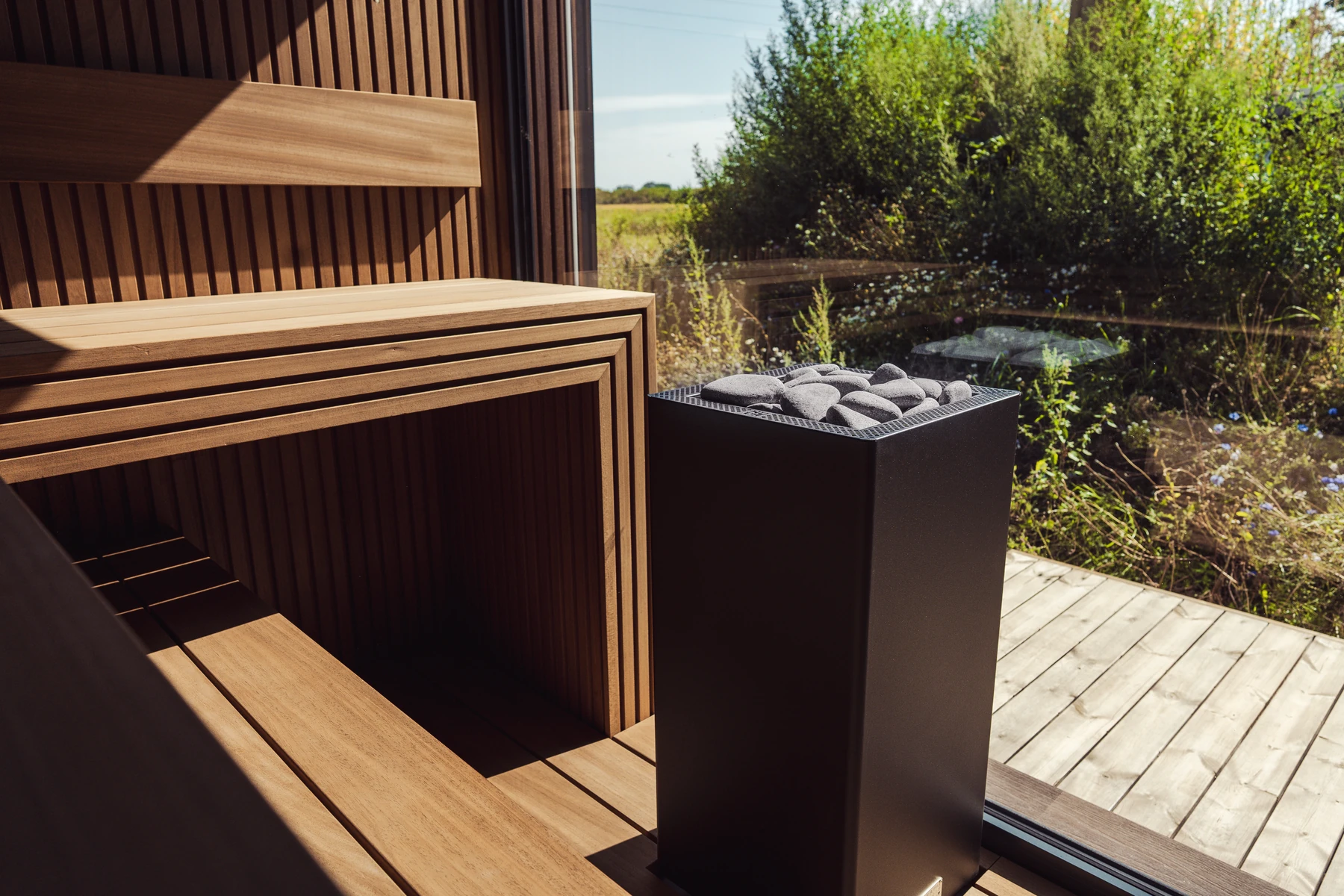HUUM CORE Black heater next to a sauna bench with a garden view through the glass