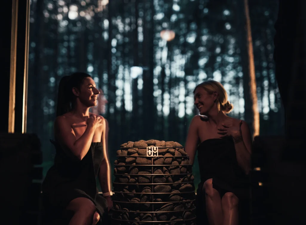 Two women relaxing in a forest view sauna beside the HUUM HIVE Mini heater