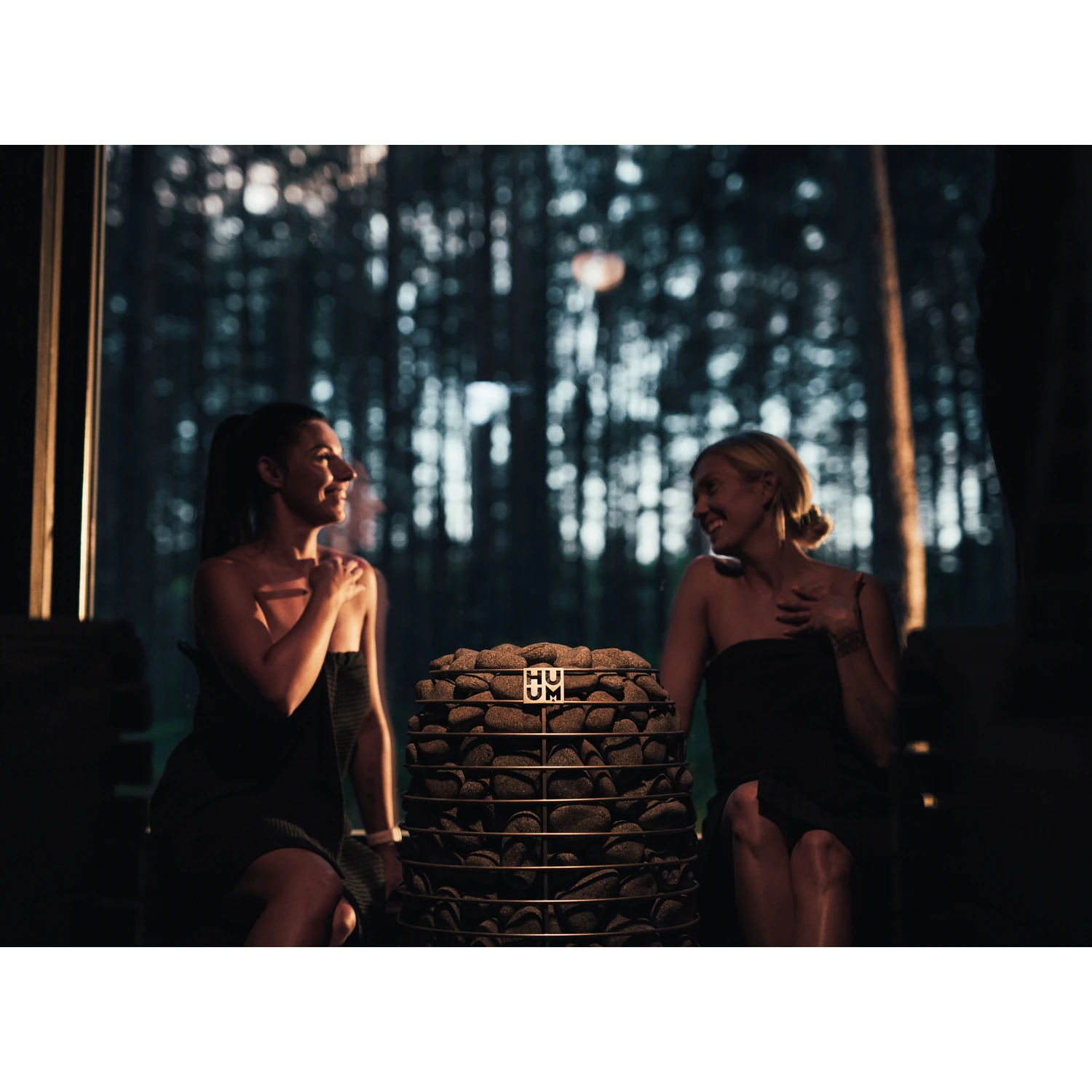 Two women relaxing in a forest view sauna beside the HUUM HIVE Mini heater