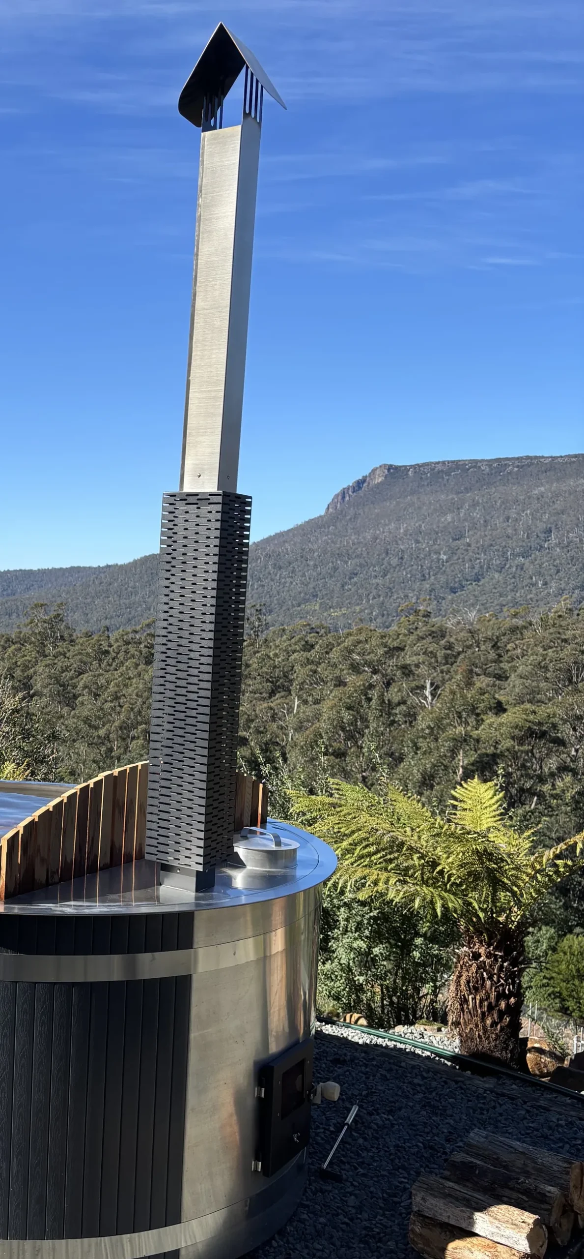 The flue and firebox door of the Roundabout hot tub, with firewood beside it and the mountain behind