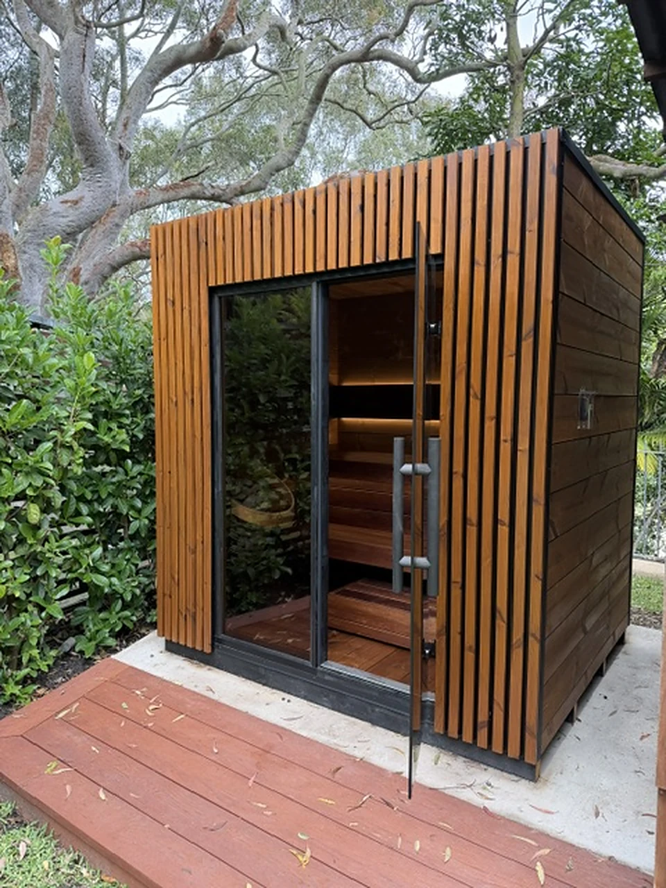 The bronze glass door and long bar handle of the Leil Viva 210 sauna, with lit benches inside and a timber deck in front